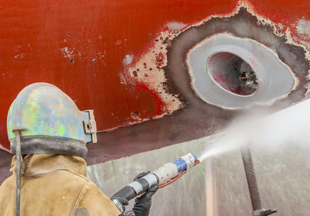 A worker using abrasive blasting equipment on a corroded boat hull